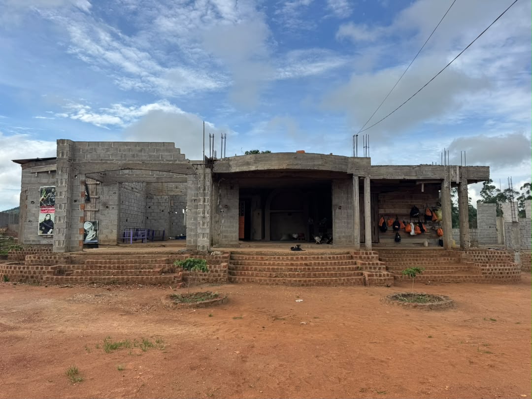 Fachada do bloco principal em obra: paredes de bloco aparente, laje plana e escadas de tijolo sobre a terra vermelha.